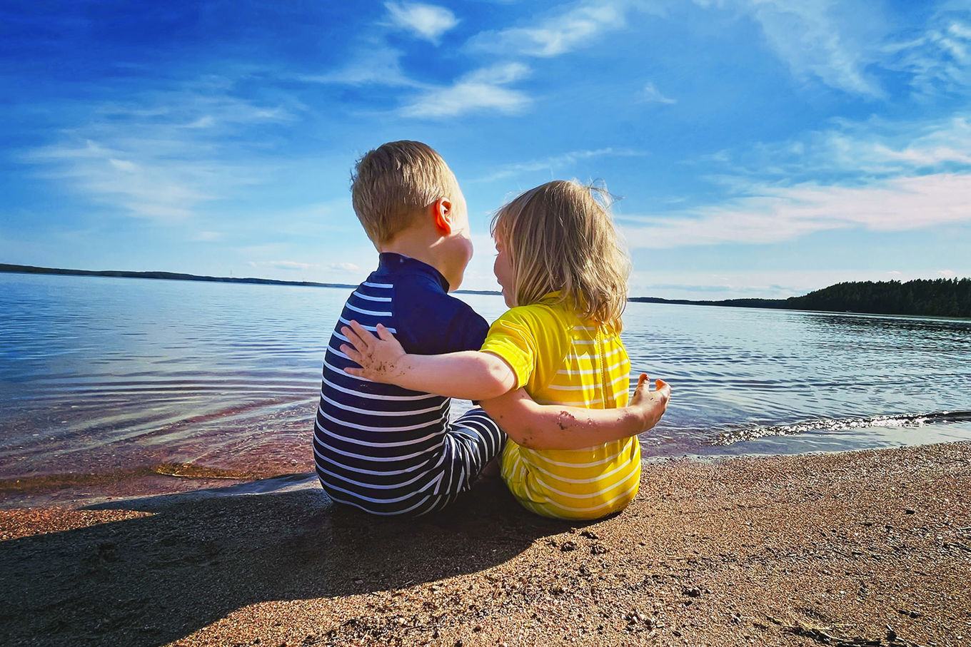 Kaksi lasta istuu hiekkarannalla ja pitää toisistaan kiinni.