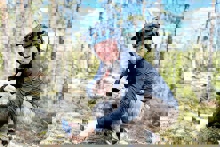 Mies kyykyssä metsäkalliolla. Mies näyttää kameralle peukkua.