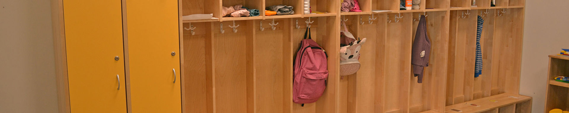 Keltainen naulakko päiväkodin aulassa. Naulakossa roikkuu lasten vaatteita ja reppuja.