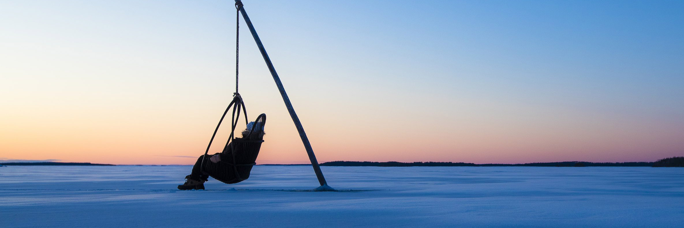 Talvinen maisema sinisen sävyissä. Ihminen istuu järven jäällä riippukeinussa.
