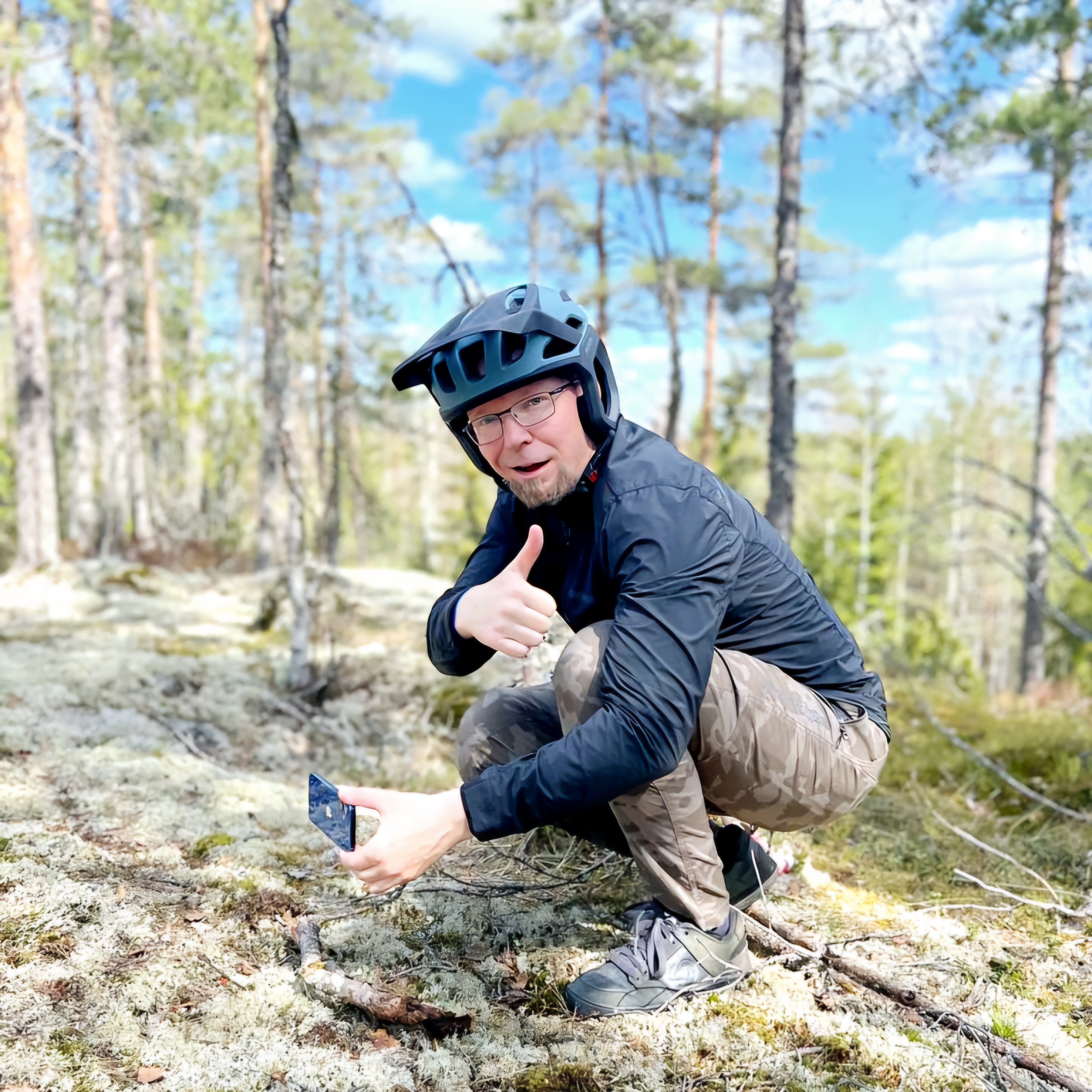 Mies kyykyssä metsäkalliolla. Mies näyttää kameralle peukkua.